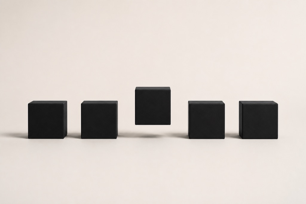 Five identical matte black cubes in a precise row on a pale bone backdrop, one lifted above the line