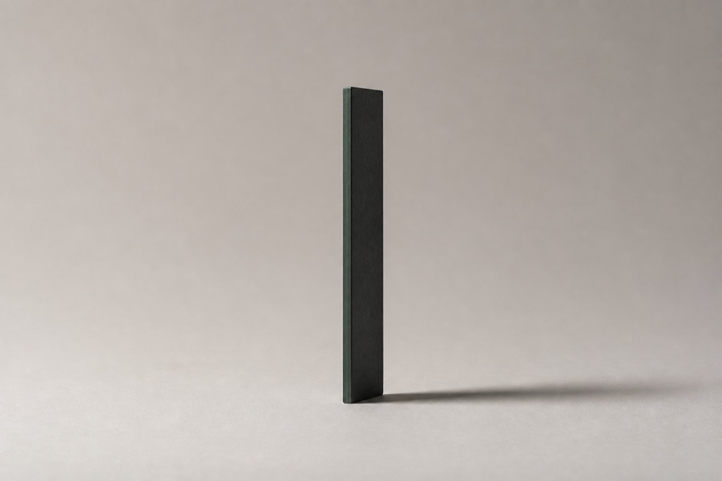 A machined slab of dark graphite standing upright on pale grey paper like a text cursor, one edge lit deep green