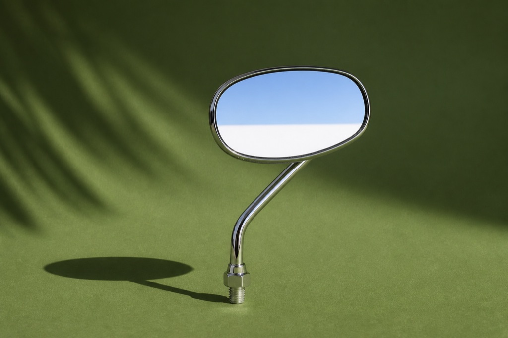 A single chrome motorcycle wing mirror standing on a palm-green backdrop, reflecting a strip of pale sky